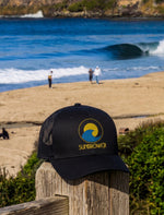 A black trucker-style cap with a yellow and blue wave 'SUNBREAKER' logo sits on a wooden post in the foreground, with a beach, ocean waves, and distant people blurred in the background.