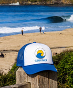 A blue and white trucker hat with a 'SUNBREAKER' logo is displayed on a wooden post in the foreground, with a beach and ocean waves in the background.