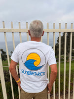 A man viewed from behind wearing a white t-shirt with a large graphic on the back featuring a yellow sun and blue wave 'SUNBREAKER' logo. He is standing in front of a white metal fence overlooking a grassy landscape and the ocean under a cloudy sky.