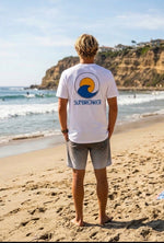 A man standing on a sandy beach facing the ocean, wearing a white t-shirt with a blue and yellow wave 'SUNBREAKER' logo printed on the back, paired with gray shorts.