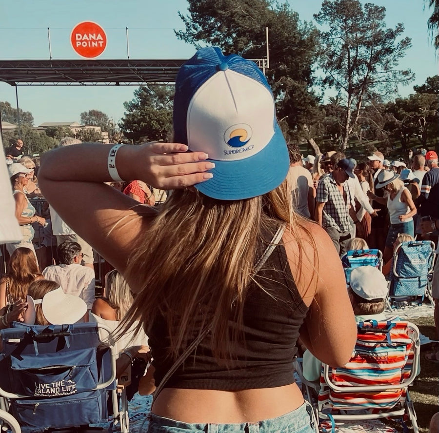 A woman seen from behind at an outdoor concert or festival, wearing a blue and white trucker-style cap with a visible 'Seychelles' logo. The background shows a crowd, chairs, trees, and a stage, with a red 'DANA POINT' sign overhead.