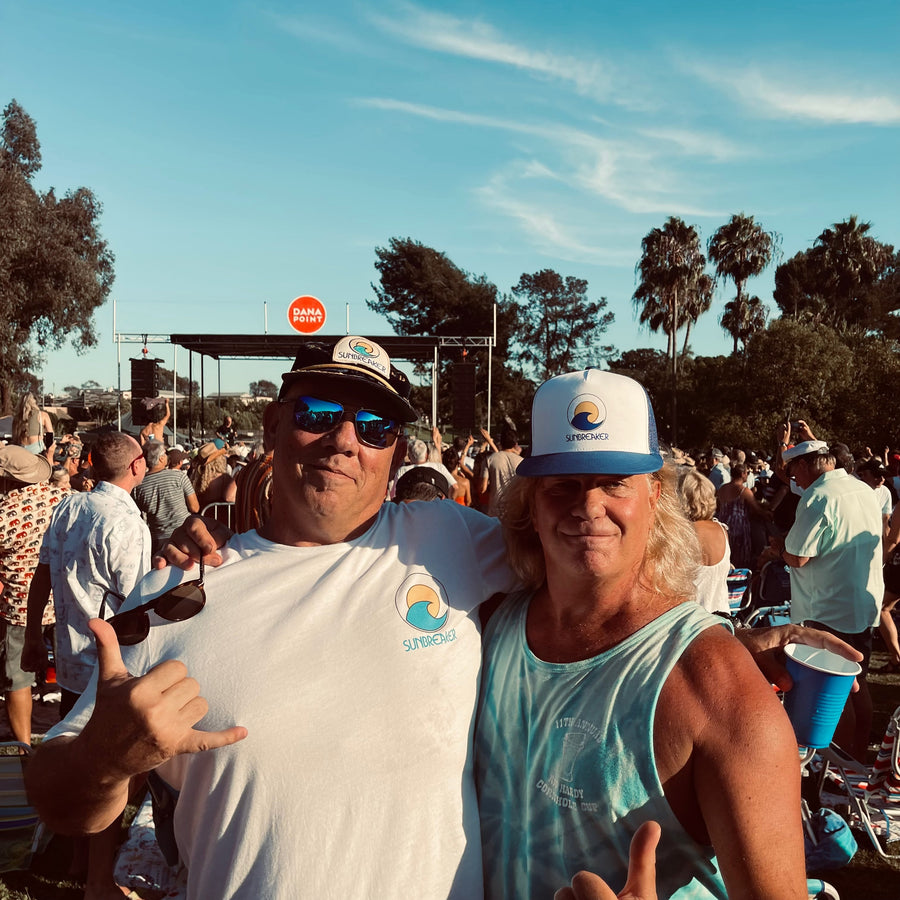 Two men posing together at an outdoor beach festival under a blue sky. The man on the left wears a white t-shirt with a blue wave graphic and a dark cap, while the man on the right wears a light blue tank top and a white cap with a blue brim.