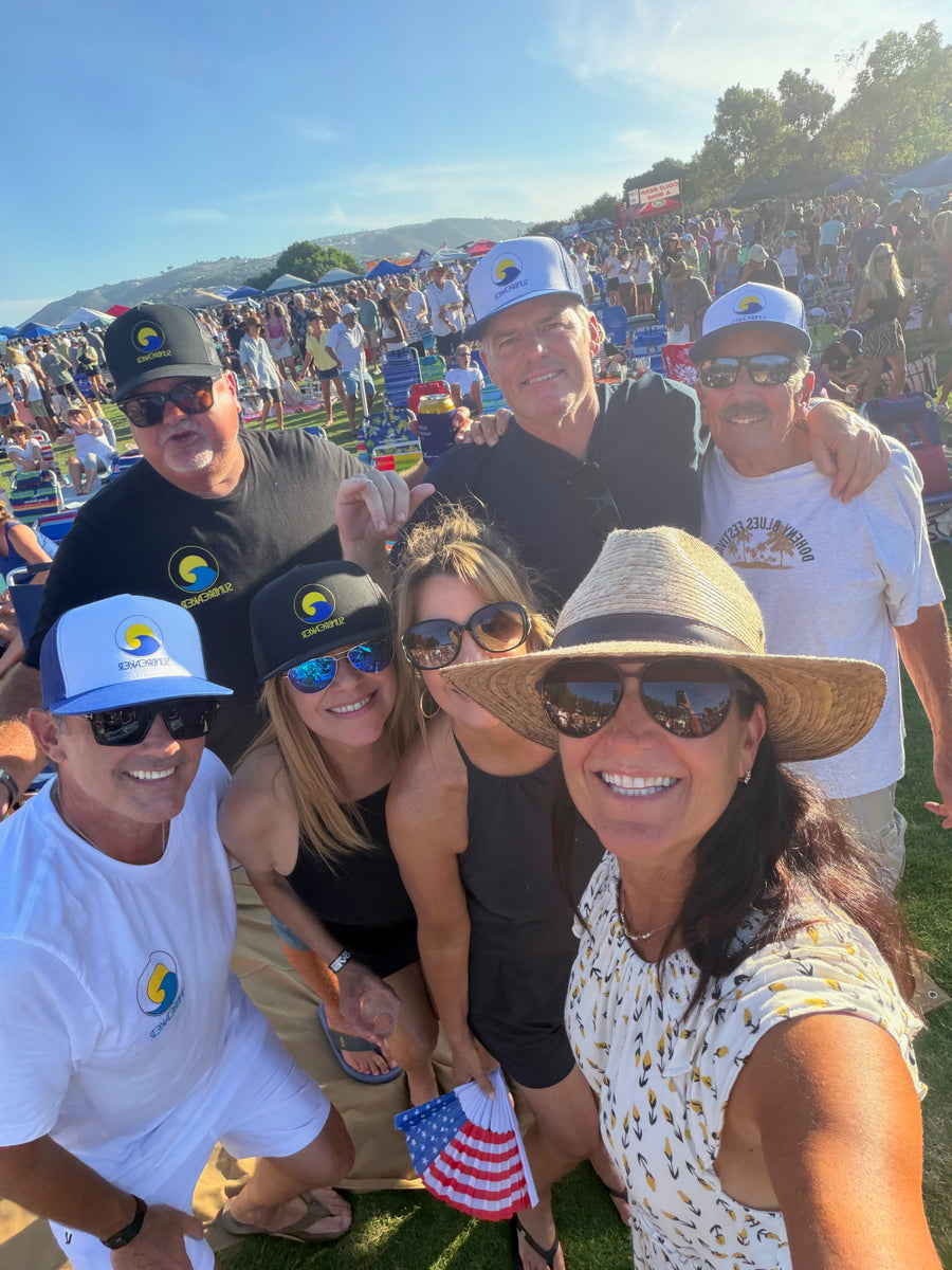 A group of seven people posing together outdoors at a large crowd event under a blue sky. Several people are wearing matching white and blue caps with a yellow crescent logo, along with matching white t-shirts featuring the same logo.