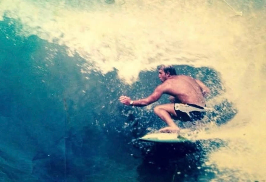 A shirtless surfer crouches on a surfboard inside a breaking wave, surrounded by white spray and blue-green water. The photo has a vintage, slightly grainy, yellowed tone.