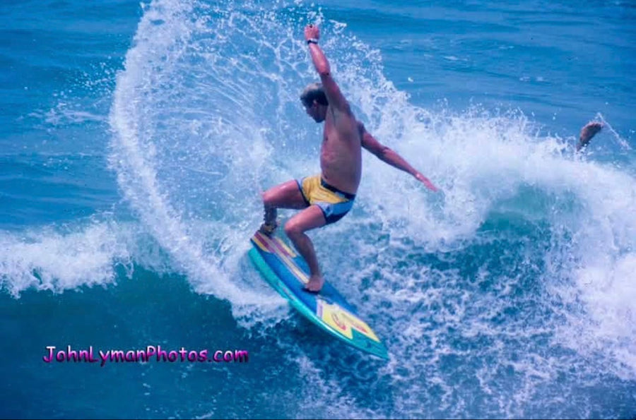 A man surfing on a blue and yellow board on a turquoise ocean wave, with white spray around him and a visible JohnLymanPhotos.com watermark in the lower left.