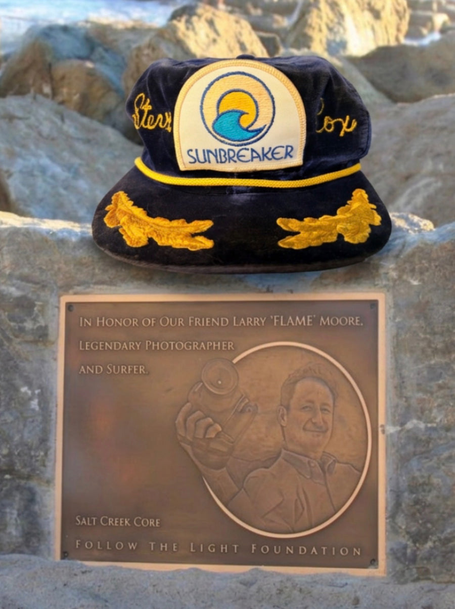 A navy blue embroidered cap with gold and white details sits on a stone surface above a bronze memorial plaque. The cap has 'SUNBREAKER' and gold script embroidery, and the plaque reads 'In Honor of Our Friend Larry 'Flame Moore,' Legendary Photographer and Surfer,' with 'Salt Creek Core' and 'Follow the Light Foundation' at the bottom.
