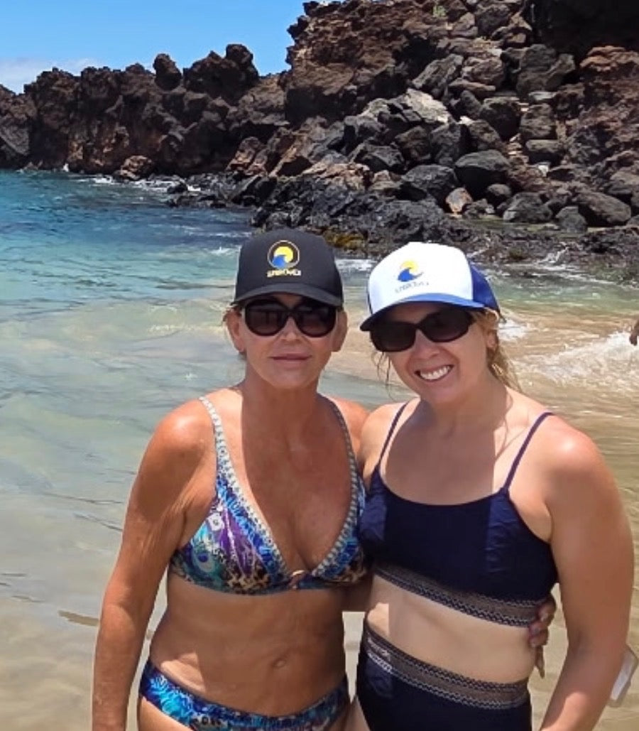 Two women posing in swimwear at a beach with rocky shoreline and blue water in the background. One wears a black cap with a visible 'SUNBREAKER' logo, and the other wears a white cap with a blue logo at Kāʻanapali Beach in Maui, Hawaii.