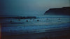 Video of people waiting in the ocean at dawn for a wave, a man surfing in the swell of a wave, two men walking on the beach carrying their surfboards under their arms, a surfboard lying on the beach with waves gently swelling up around it and 6 people standing in front of the ocean with their surfboards under their arms at daybreak watching the sunrise.