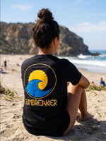 A woman sitting on a sandy beach, viewed from behind, wearing a black t-shirt with a large yellow and blue wave 'SUNBREAKER' logo printed on the back.