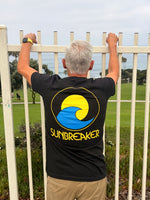 A man viewed from behind wearing a black t-shirt with a large yellow and blue 'SUNBREAKER' logo and text on the back, leaning on a white fence with a golf course and ocean in the background.