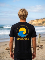 A man standing on a beach, viewed from behind, wearing a black t-shirt with a large yellow and blue wave 'SUNBREAKER' logo printed on the back.