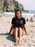 A woman sitting on a sandy beach wearing a black t-shirt with a yellow and blue wave 'SUNBREAKER' logo printed on the chest. The background features a rocky coastline, ocean waves, and other beachgoers.