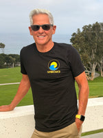 A smiling man with silver hair and sunglasses wearing a black t-shirt with a yellow and blue 'SUNBREAKER' logo, standing outdoors on a white wall with a golf course and ocean in the background.