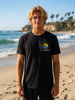 A man standing on a sandy beach wearing a black crewneck t-shirt with a yellow and blue wave 'SUNBREAKER' logo printed on the left chest.