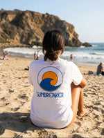 A person sitting on a sandy beach with their back to the camera, wearing a white t-shirt with a blue and orange wave 'SUNBREAKER' logo printed on the back. The background features a rocky coastline and ocean waves.