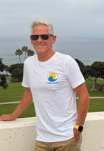 A smiling man with white hair and sunglasses stands outdoors by a concrete ledge overlooking a grassy park and ocean, wearing a white t-shirt with a blue and yellow 'SUNBREAKER' logo on the chest.