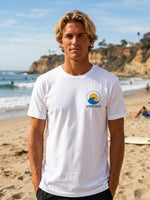 A man standing on a sandy beach wearing a white crewneck t-shirt with a yellow and blue wave 'SUNBREAKER' logo on the left chest.