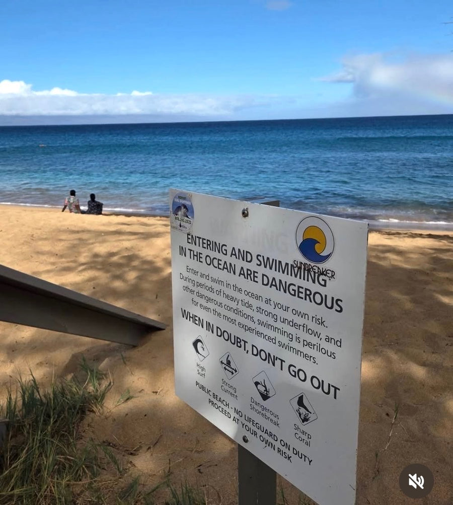 A beach warning sign in the foreground reading 'Entering and Swimming in the Ocean are Dangerous' with ocean and sand in the background.  The sign has a 'SUNBREAKER' sticker in the upper right corner.